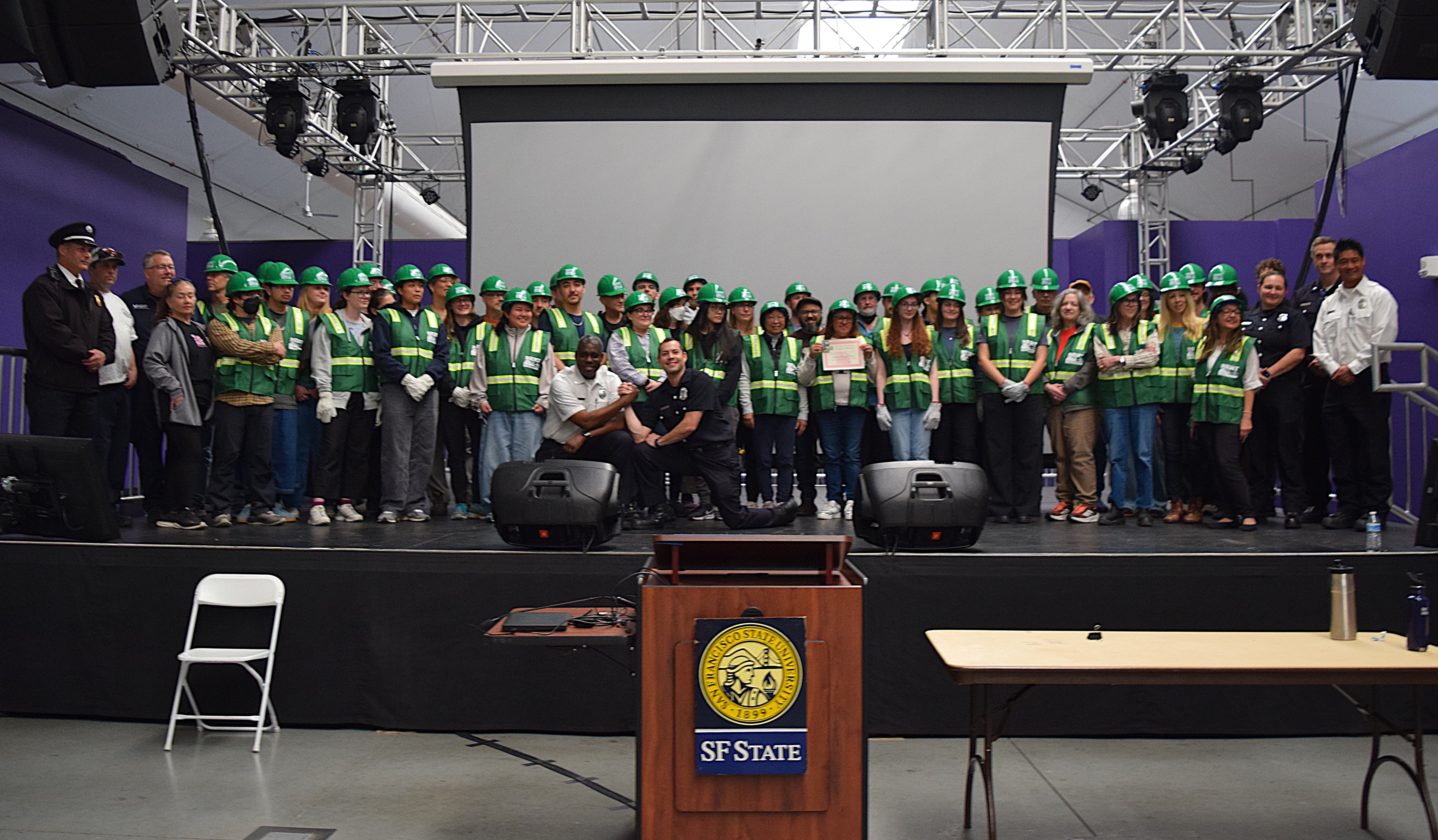 A large group on a stage in front of a SF State podium poses for a picture with NERT vests and hard hats