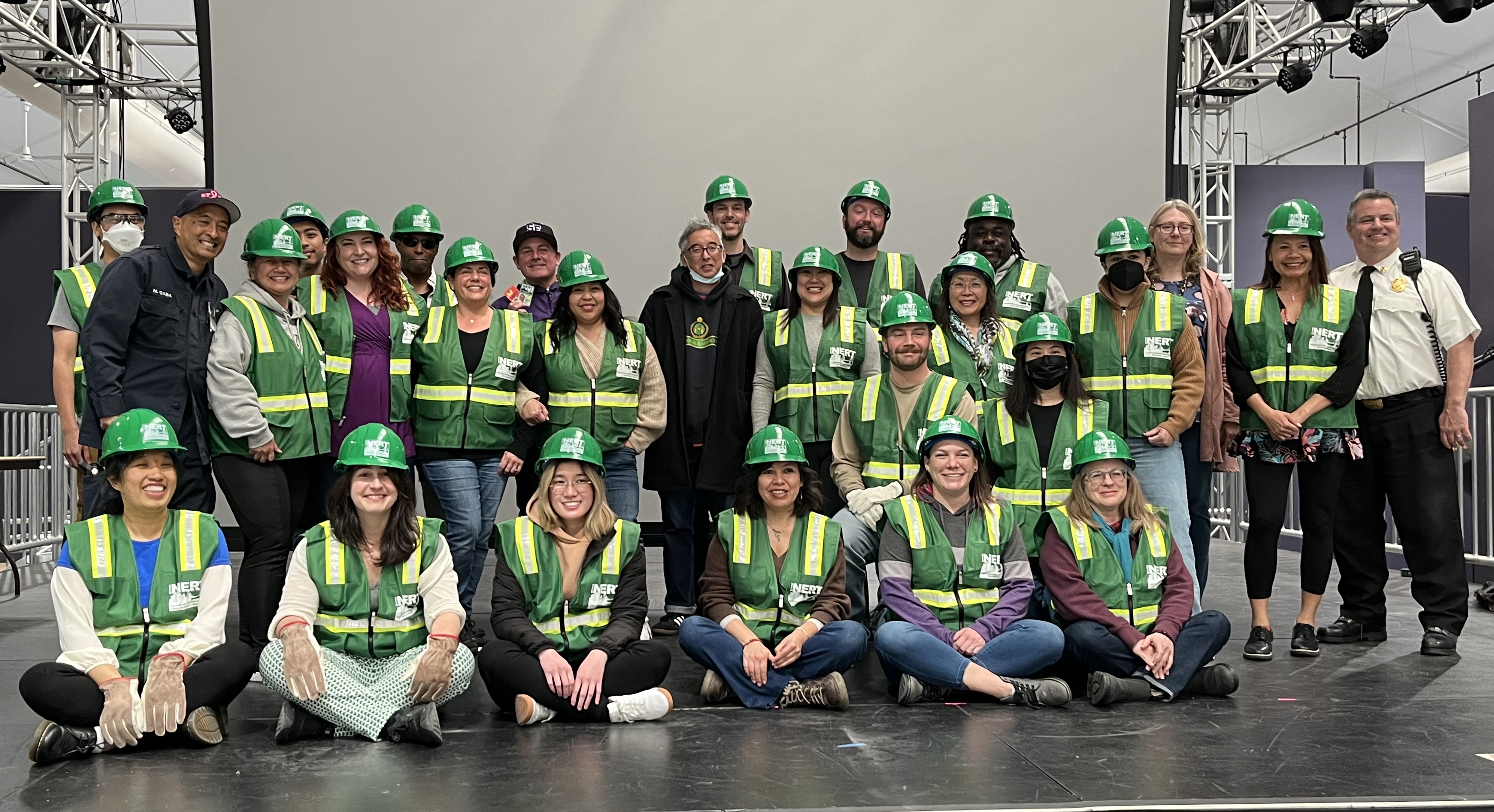 a group of recently certified NERT members in green and yellow vests and green hard hats pose for a group picture on a stage at SF state with some people sitting in front and some standing in back